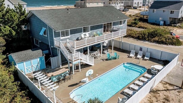 Uma casa de dois andares com varanda para uma praia, piscina e área de estar externa.
