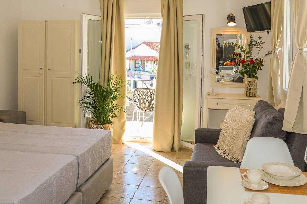 Habitación de hotel con cama, sofá, mesa de comedor con platos, planta en maceta y balcón con vistas a edificios.