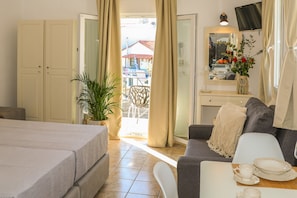 Ein Hotelzimmer mit Bett, einer Couch, einem Esstisch mit Tellern, einer Topfpflanze und einem Balkon mit Blick auf Gebäude.