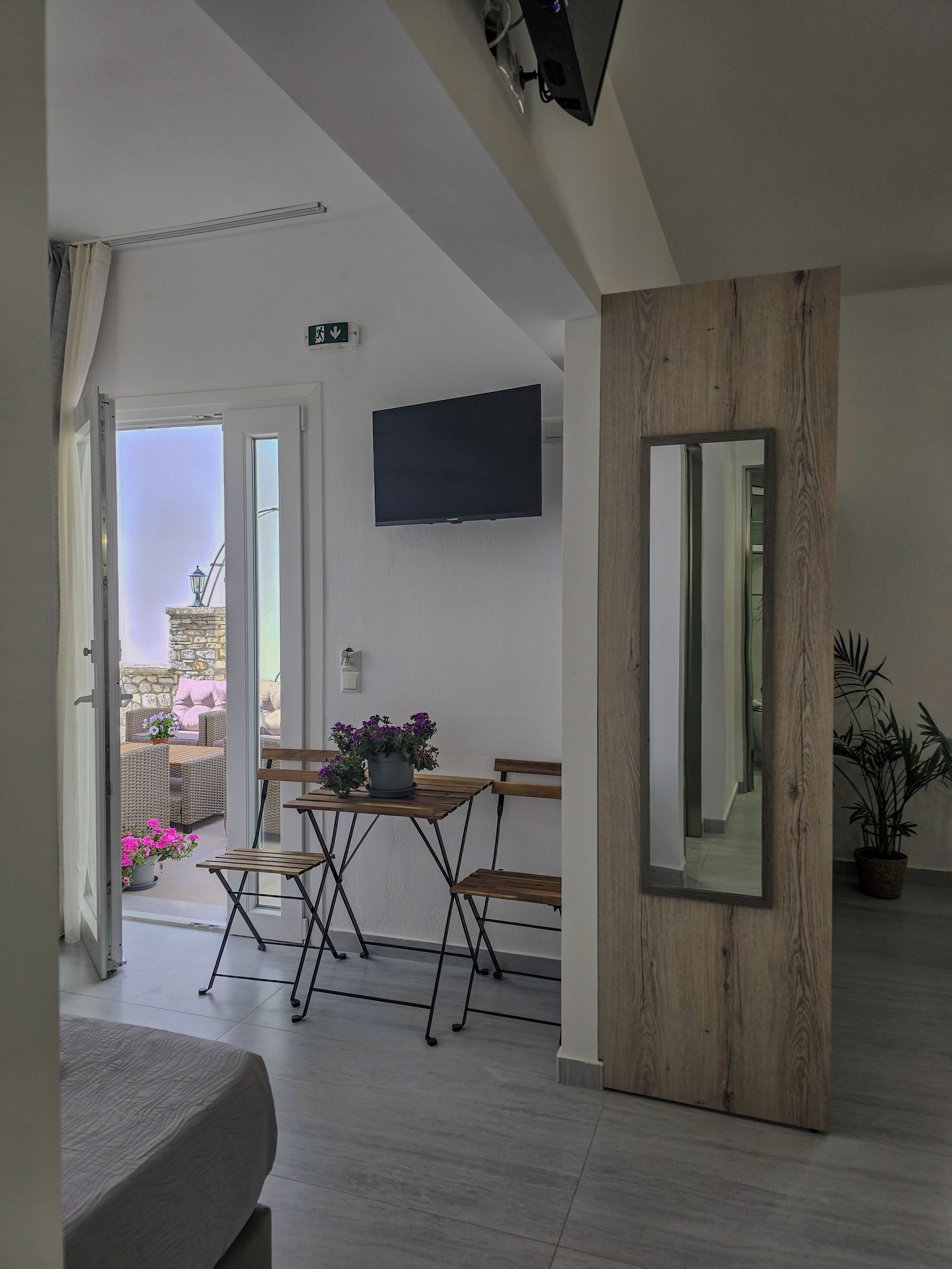 A modern room with a flat-screen TV, a wooden accent wall, a dining area with a table and chairs, and a view through an open door to an outdoor patio.