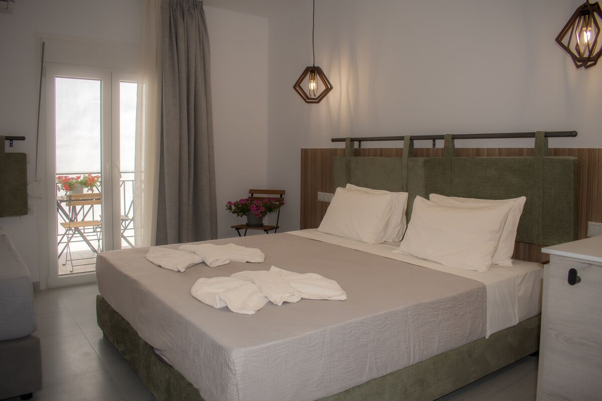 Una cama bien hecha con ropa y toallas blancas, un cabecero de madera, una mesita de noche con una lámpara y vistas al exterior a través de una puerta de cristal.