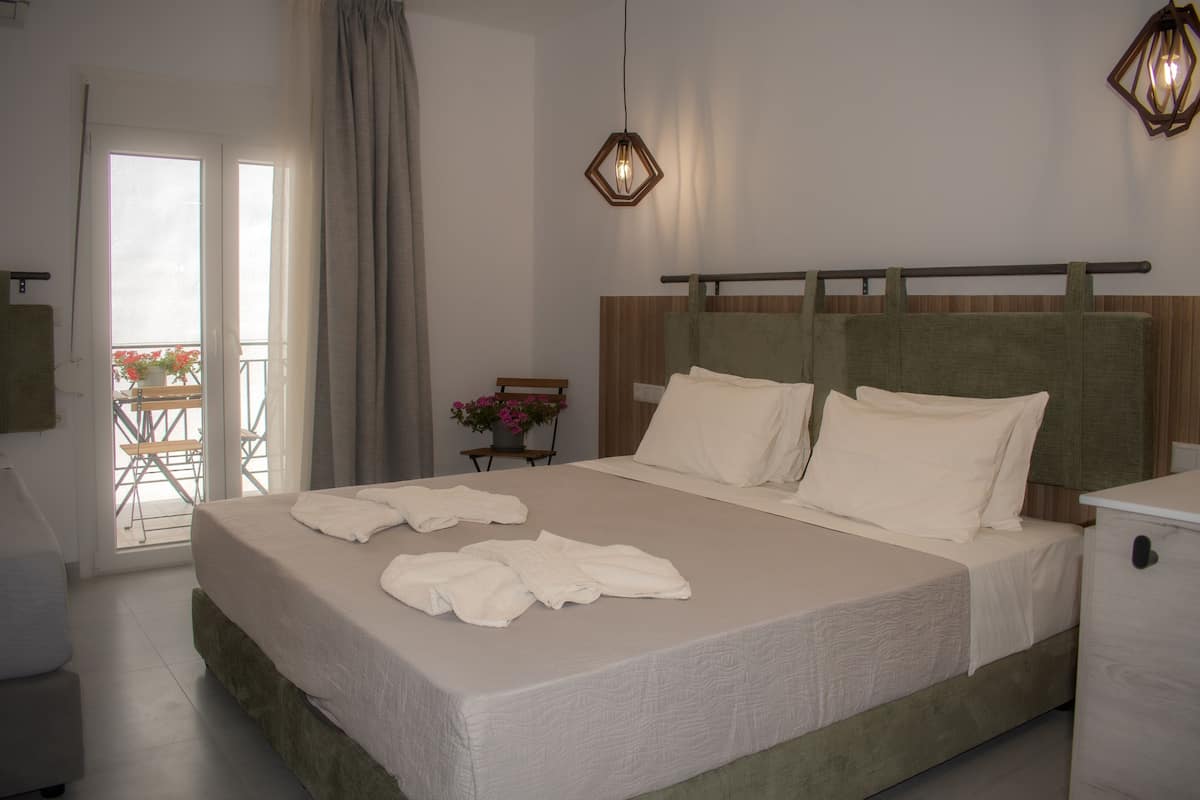 Una cama bien hecha con ropa y toallas blancas, un cabecero de madera, una mesita de noche con una lámpara y vistas al exterior a través de una puerta de cristal.