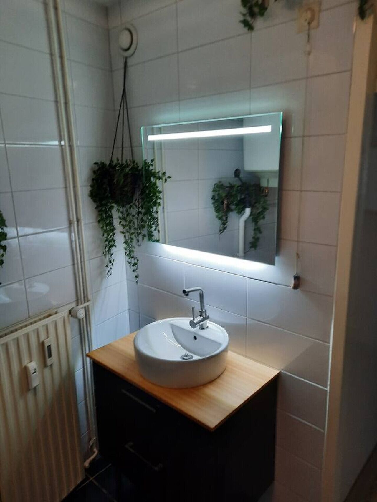Une salle de bain avec un mur recouvert de carreaux blancs, un meuble-lavabo en bois, un miroir et des plantes suspendues.
