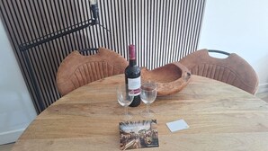 A round wooden table with a bottle of wine and two glasses, a placemat with a welcome message, and a chair with a woven seat.