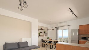 A modern living room with a grey sofa, a wooden coffee table, and a dining area with a wooden table and chairs. There is a kitchen area with wooden cabinets and a refrigerator. - Perivoli SeaView Cottage, a Rural Bliss of Nature, Close to tavernas & beach (Rethymno)