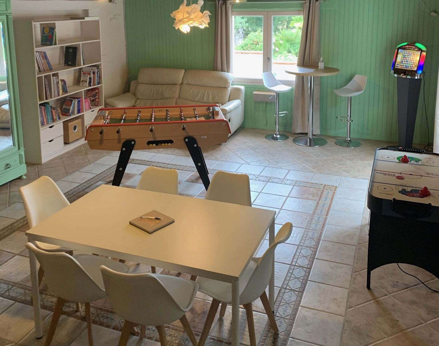 Een gezellige kamer met een tafelvoetbaltafel, een bank, een eettafel met stoelen, een jukebox en een boekenkast.