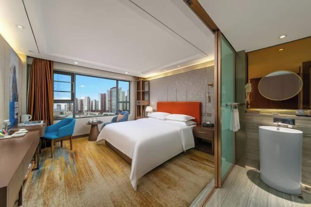 Ein Hotelzimmer mit Bett, Schreibtisch, Stuhl und einem großen Fenster mit Blick auf die Stadt.