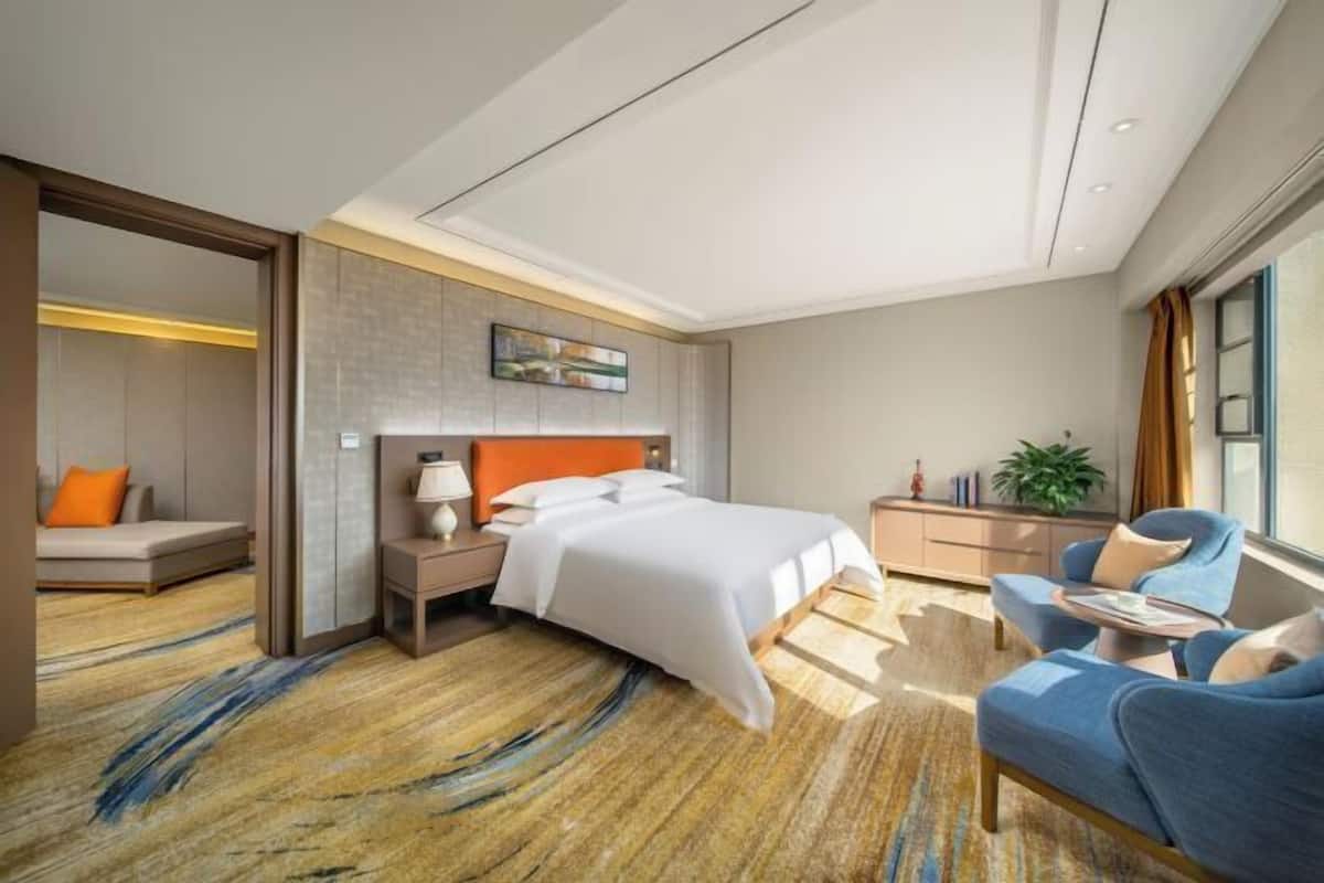Ein Hotelzimmer mit Bett, Nachttisch, Stuhl, Schreibtisch, Pflanze und Fenster.