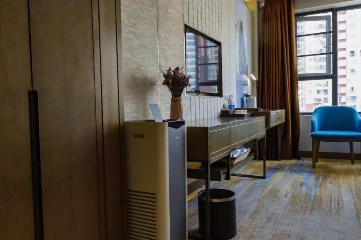 A hotel room with a large window, a desk with a vase of flowers, a blue chair, and a trash bin.