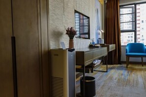 A hotel room with a large window, a desk with a vase of flowers, a blue chair, and a trash bin.