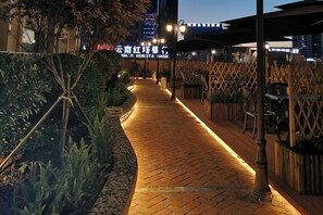 Un chemin pavé de briques, bordé de rampes en bois et agrémenté de plantes, mène à un bâtiment dont l’enseigne est illuminée.