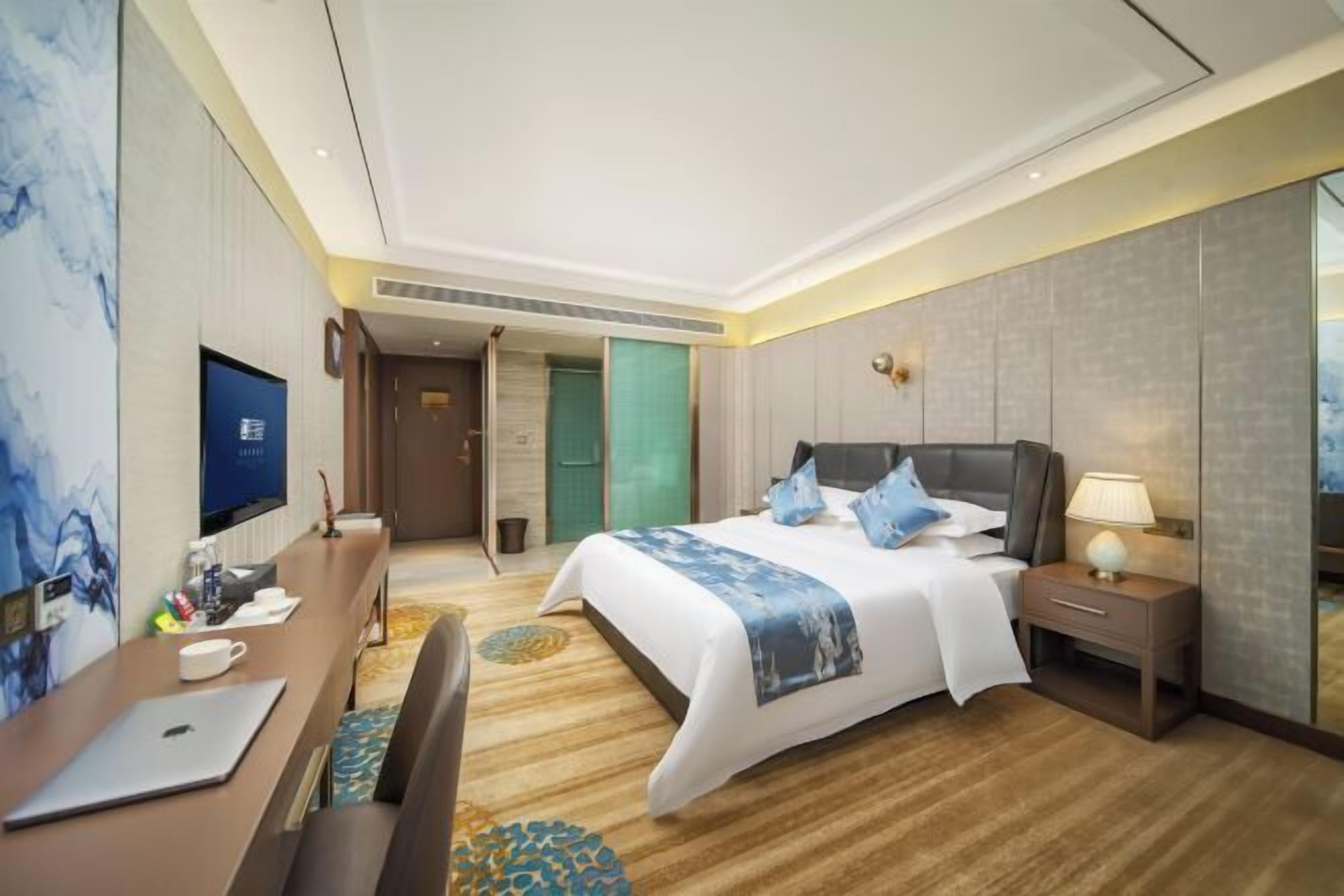A modern hotel room with a large bed, a desk with a laptop, a TV, and a mirror.