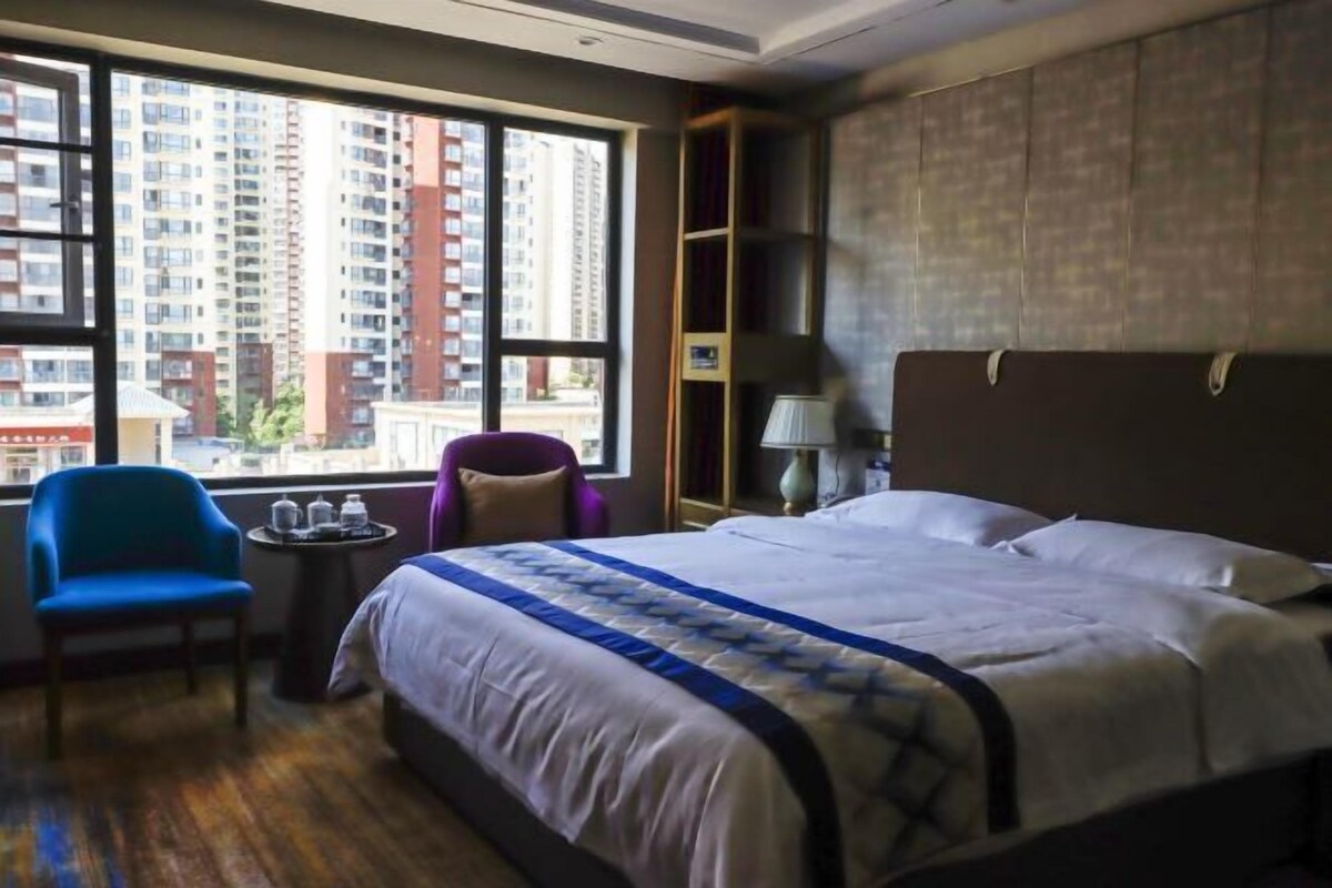 A hotel room with a large bed, a chair, a small table with a lamp, and a view of tall buildings through the window.