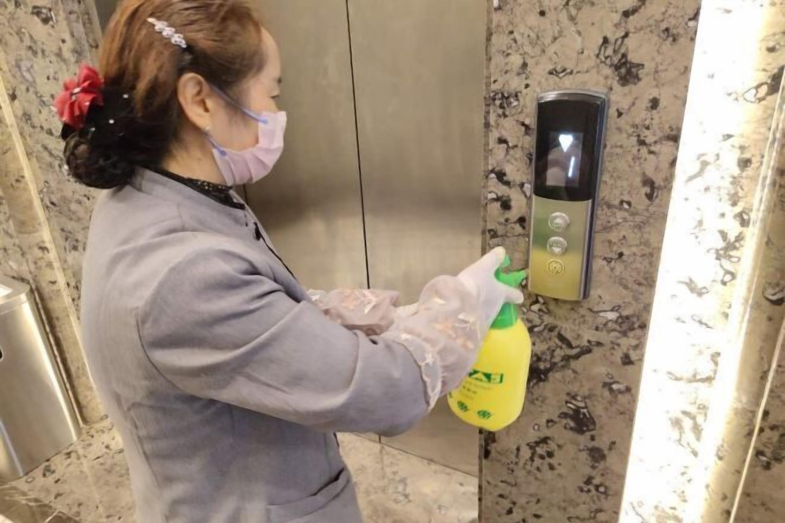 A person in a grey uniform spraying a yellow bottle on a metallic elevator control panel.