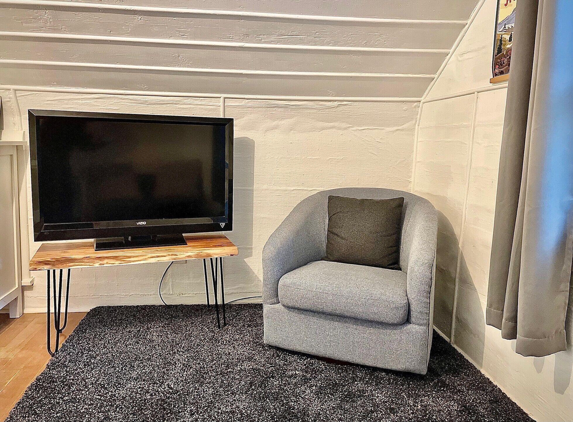 A living room with a flat-screen TV on a wooden stand, a grey armchair, and a black and white rug.