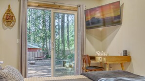 Ein Schlafzimmer mit einem Bett, einem Schreibtisch und einem Stuhl. Eine Glastür führt in einen Hinterhof mit einer Holzterrasse und einem roten Gebäude.