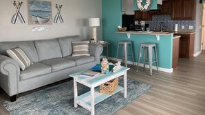 A living room with a grey sofa, a coffee table, and a kitchen area with a teal accent wall, a sign reading "Beach House," and a small island counter. - Cozy one-bedroom condo, ideal for a romantic couple's getaway. (Carolina Beach)