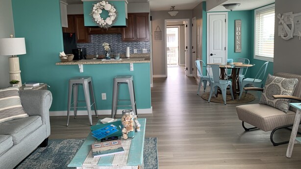 A living room with a sofa, a coffee table, and a kitchen area with a sign reading "BEACH HOUSE". - Cozy one-bedroom condo, ideal for a romantic couple's getaway. (Carolina Beach)