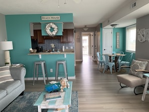 Un salon avec un canapé, une table basse et un coin cuisine agrémenté d’une pancarte indiquant « MAISON DE PLAGE ».