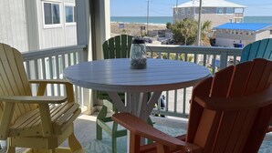 A covered outdoor seating area with a round table and Adirondack chairs, overlooking a beach and ocean. - Cozy one-bedroom condo, ideal for a romantic couple's getaway. (Carolina Beach)