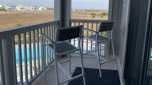 A balcony with two chairs and a view of a swimming pool and buildings in the distance. - Cozy one-bedroom condo, ideal for a romantic couple's getaway. (Carolina Beach)