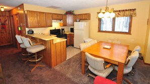 A compact kitchen with wooden cabinets, a central island with bar stools, a dining table with chairs, and a window with blinds. - Ski-in ski-out 3 Bdrm Chalet w sauna on slopes & lake - Sleeps 8 (Upson)