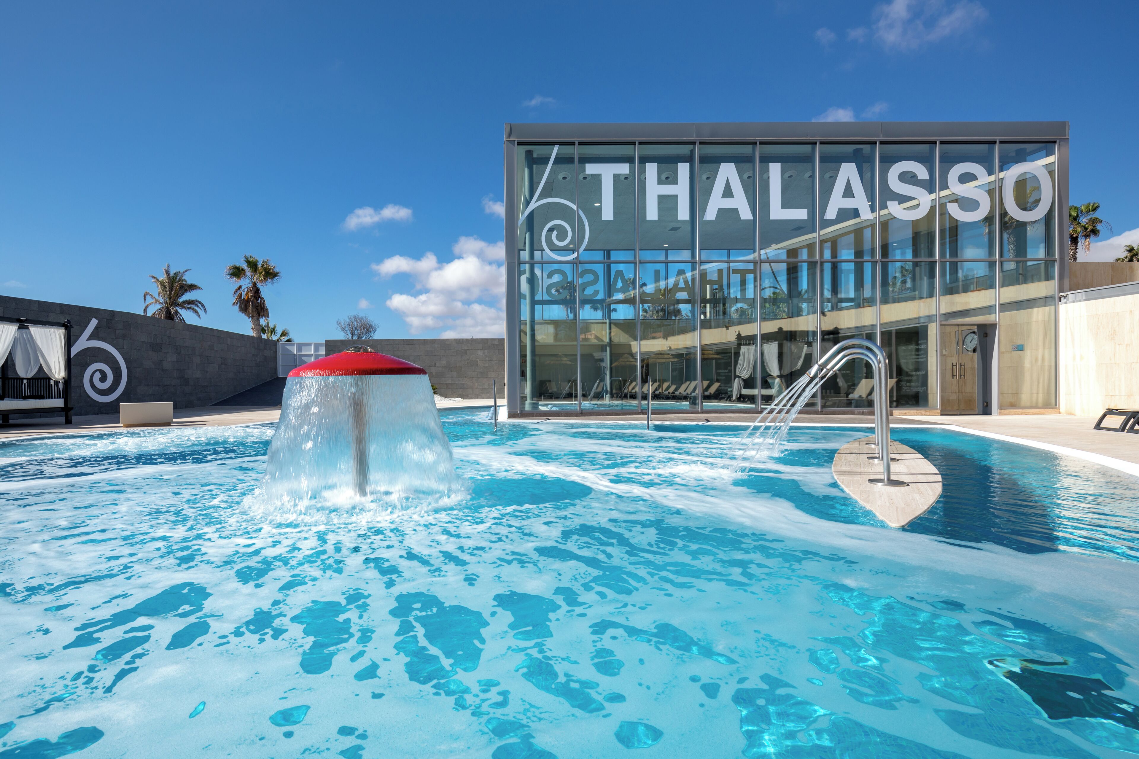 Ett modernt poolområde med en glasbyggnad i bakgrunden, där det står ”THALASSO”.