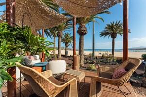 Un espacio al aire libre con mobiliario de mimbre, una palmera y vista a la playa.