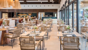 Une salle à manger spacieuse avec de hauts plafonds, de grandes fenêtres et des chaises en osier. Les tables sont dressées avec des verres à vin et des couverts.