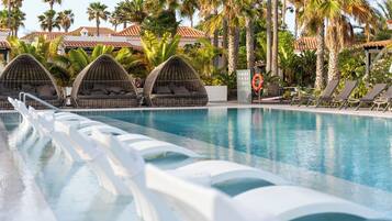 A swimming pool with lounge chairs and a covered seating area, surrounded by palm trees and a building with a tiled roof.