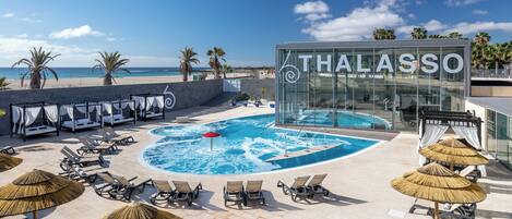 A pool area with lounge chairs and umbrellas, a glass building with 'THALASSO' signage, and a beach view.