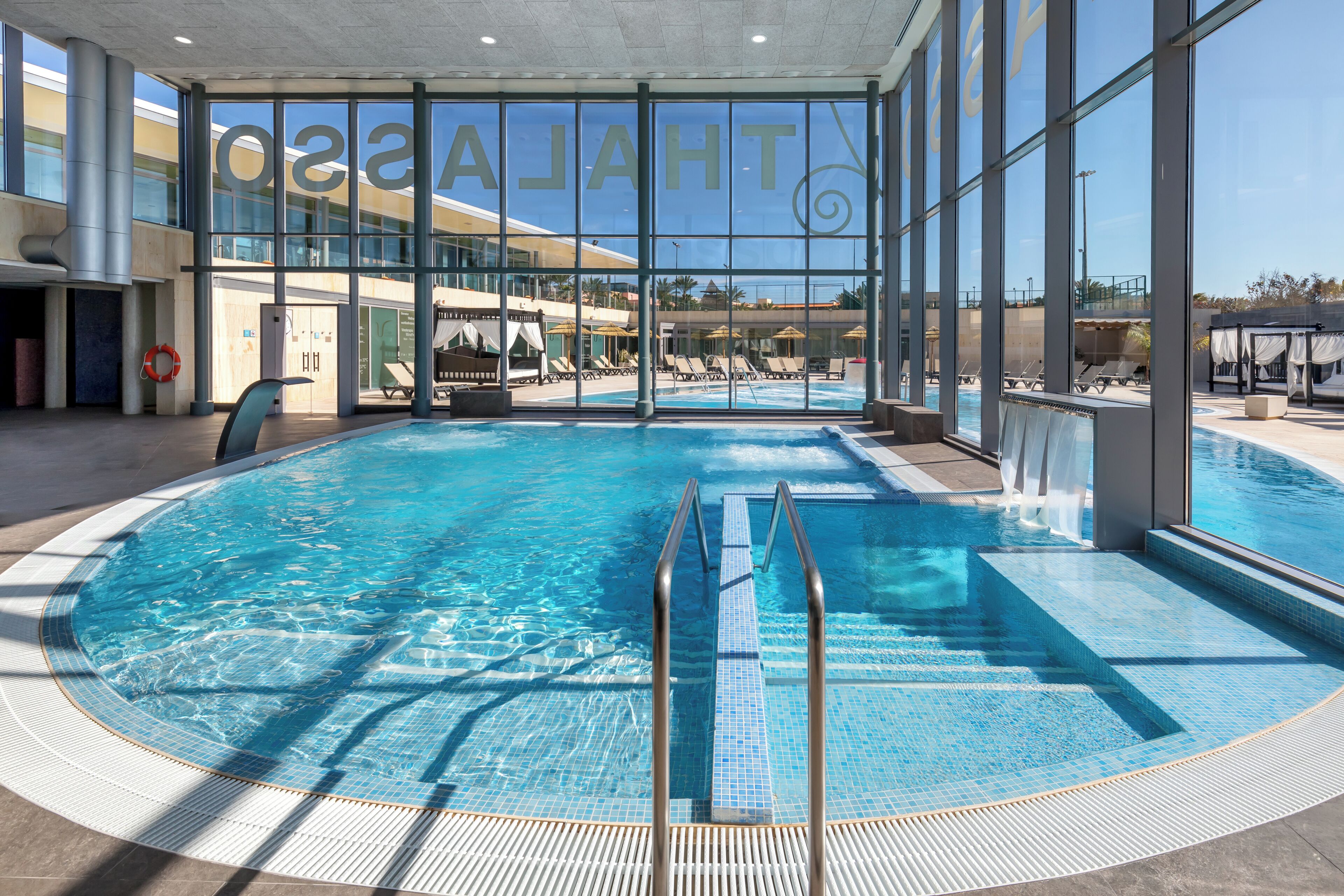 Une piscine intérieure à l’eau d’un bleu limpide, équipée d’une bouée de sauvetage et offrant une vue sur la façade de l’hôtel grâce à de grandes fenêtres.