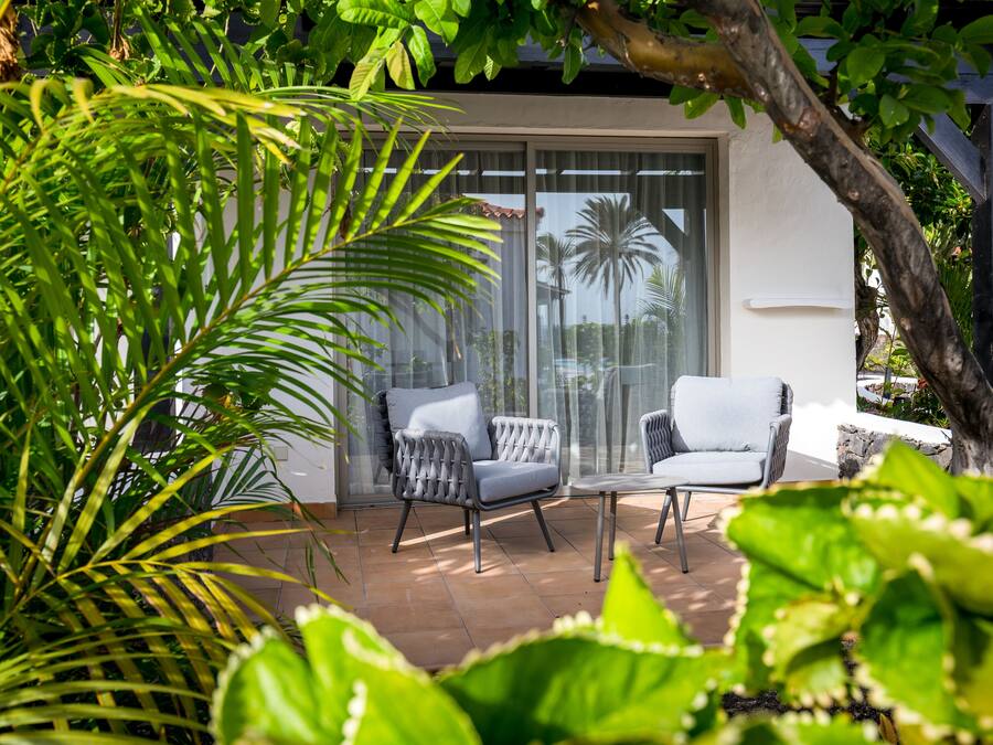 Un espace patio aménagé avec deux fauteuils gris à revêtement rembourré et une table en verre, entouré d’une végétation luxuriante et agrémenté d’une grande fenêtre donnant sur un palmier.