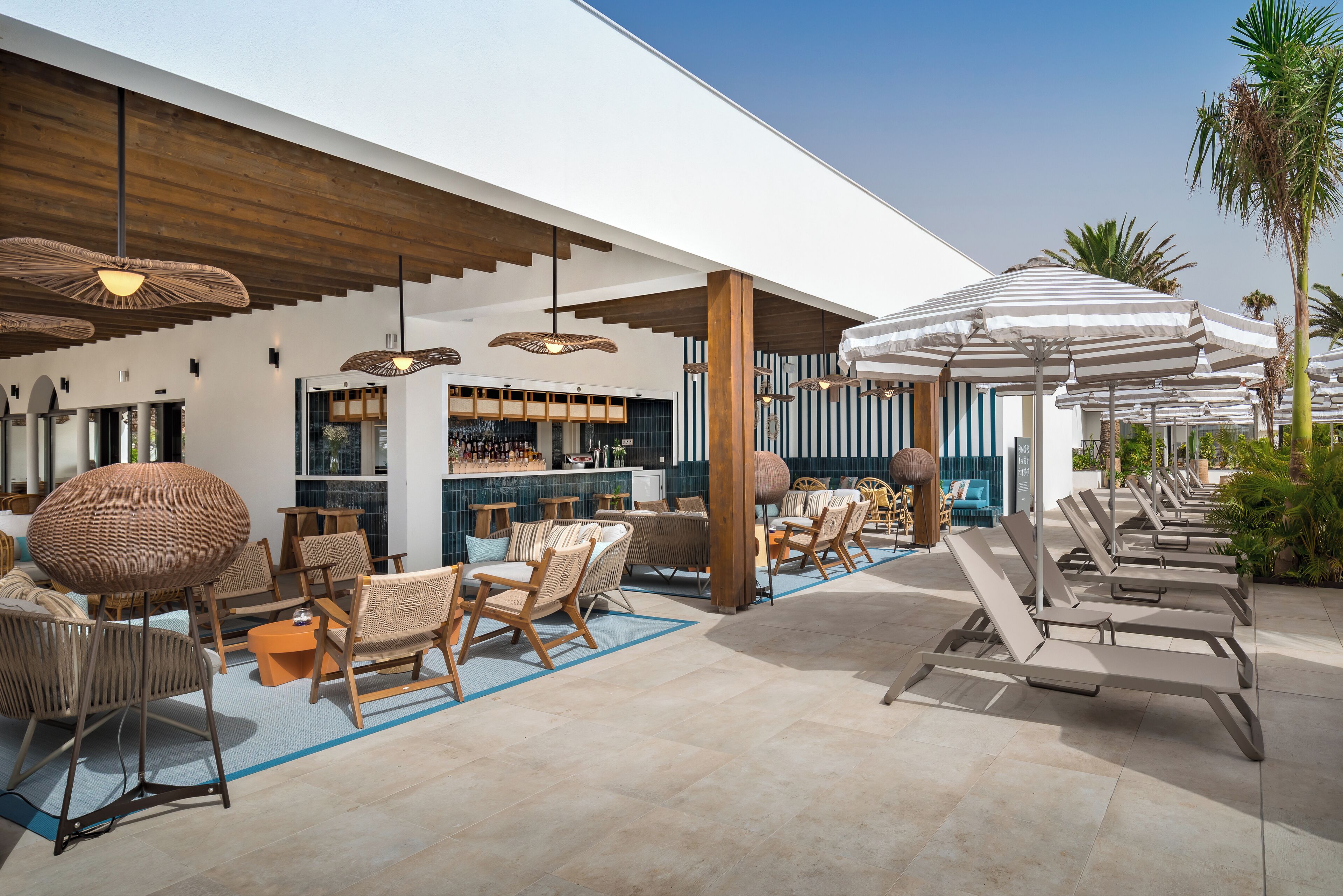 A modern outdoor lounge area with wicker furniture, striped umbrellas, and a bar with a blue-tiled backsplash.