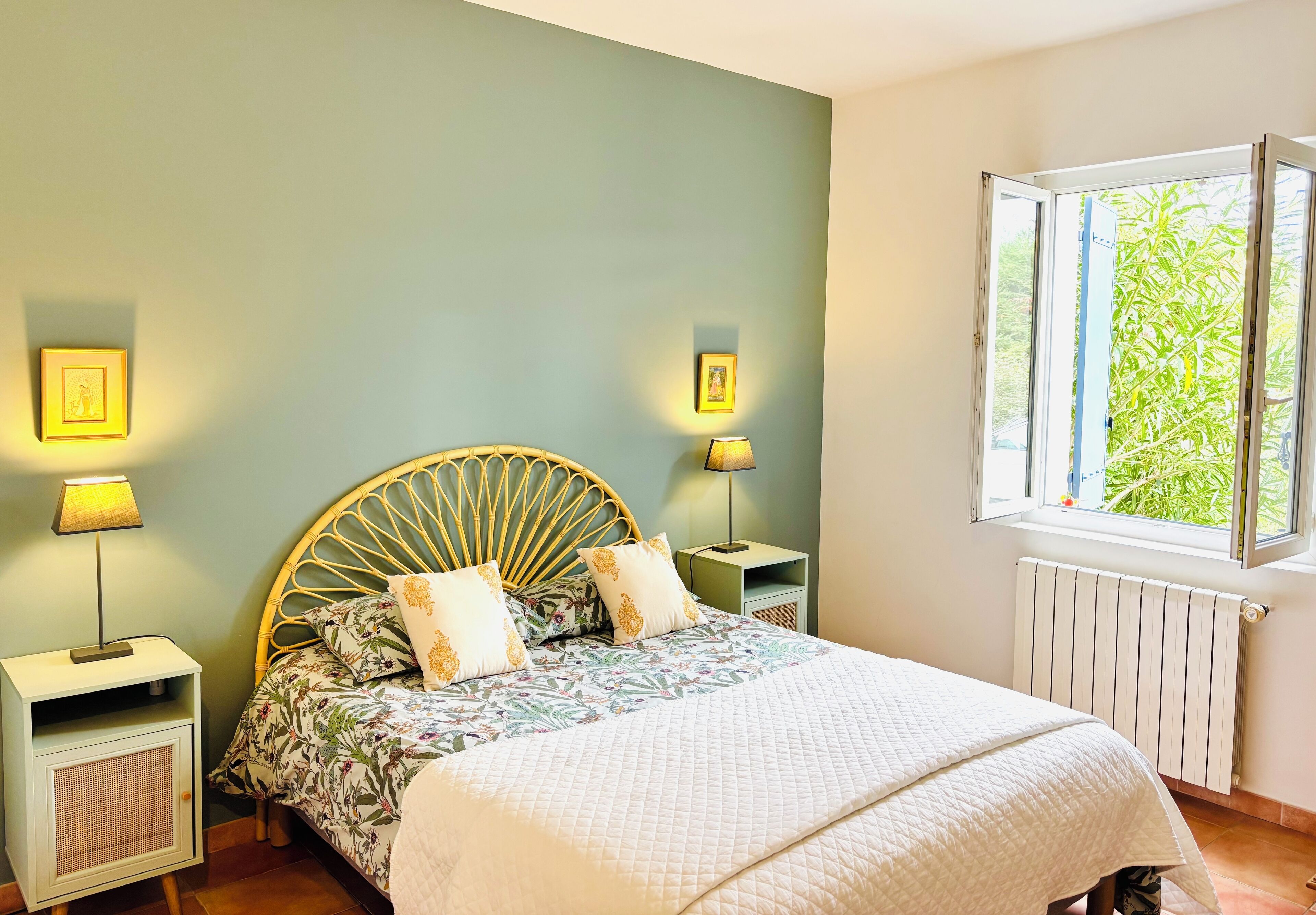 A bedroom with a bed, two bedside tables, two wall-mounted lamps, a window with a view of greenery, and a radiator.