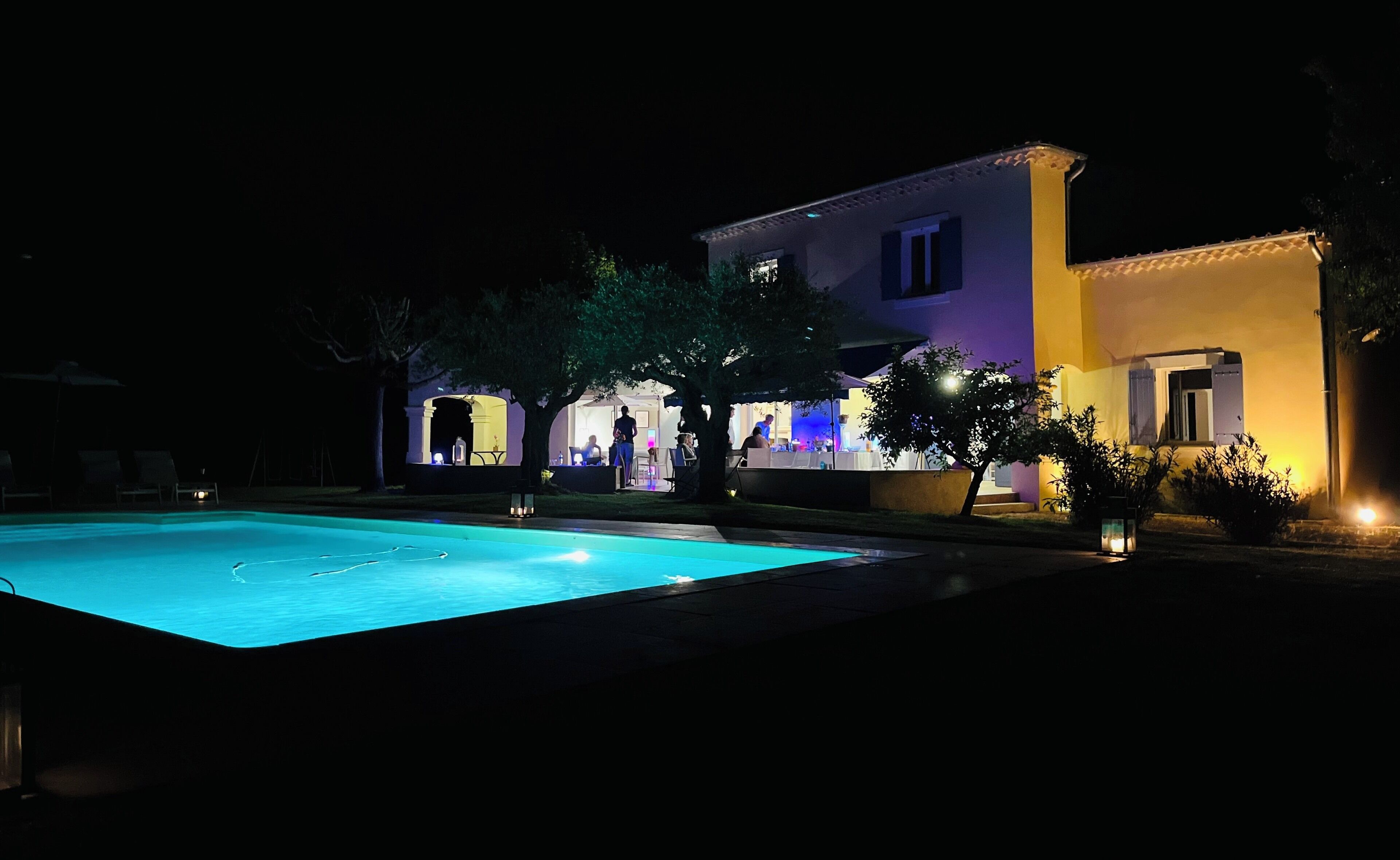 Vue sur un bâtiment avec une piscine, des espaces de détente en plein air et un patio bien éclairé, le tout vu depuis le bord de la piscine.