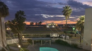A swimming pool with a view of the sunset, palm trees, and a fenced area with a white building in the background. - Little bit of Paradise (Corpus Christi)
