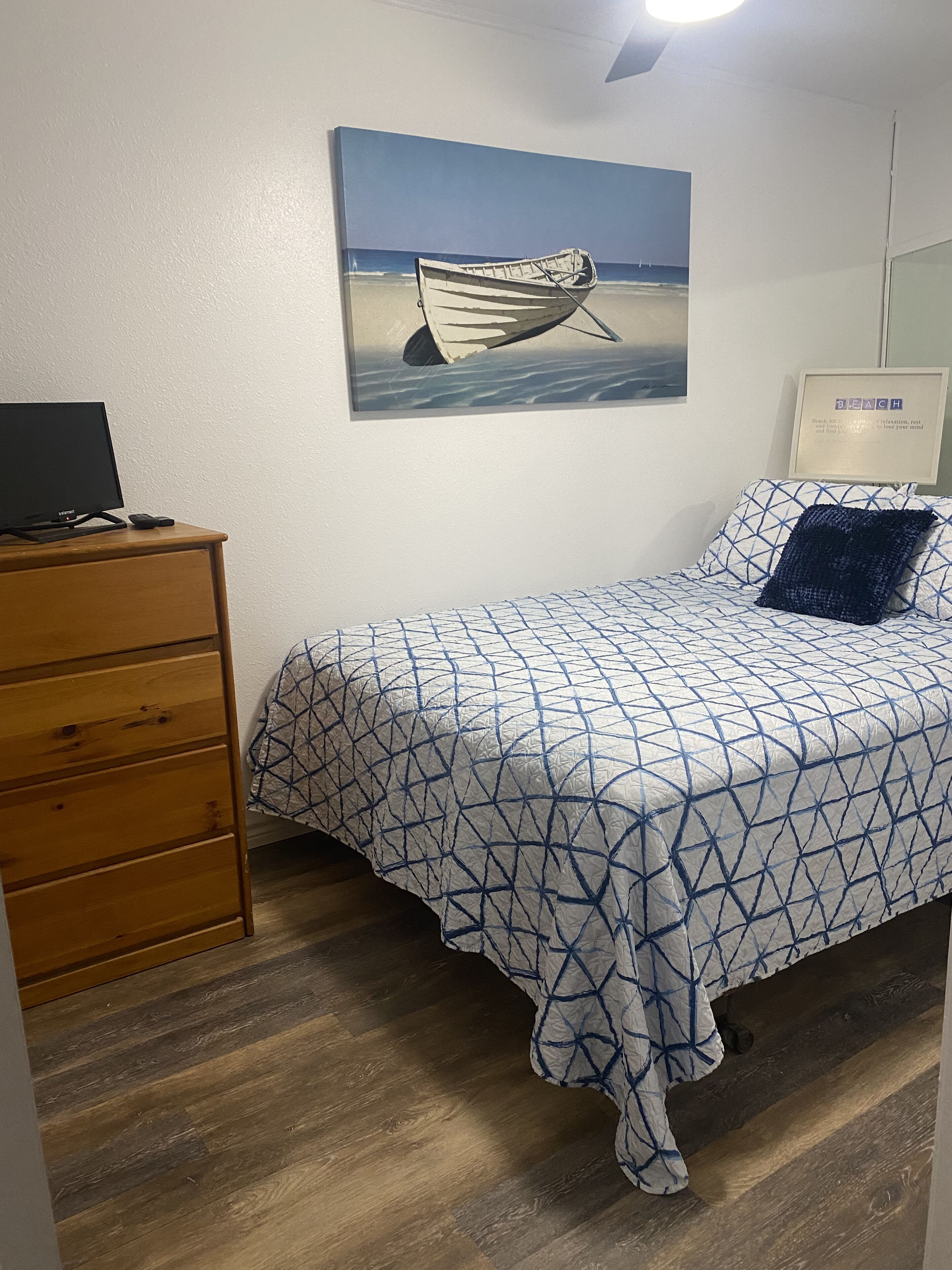 A bedroom with a bed, a dresser, a painting of a boat on the wall, and a framed sign that says "BEACH".