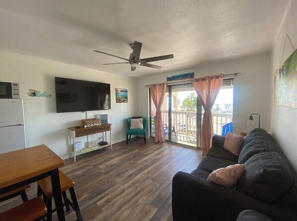 Un salon avec un plancher en bois, un canapé noir, une table en bois, un fauteuil de couleur turquoise, un ventilateur de plafond, un téléviseur fixé au mur et une fenêtre avec des rideaux.
