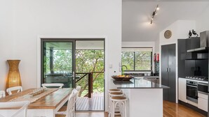 A modern kitchen with a central island, a dining area with a wooden table and white chairs, and large windows offering a view of greenery. - Mulivista - spacious home with ocean views & pool (Suffolk Park)