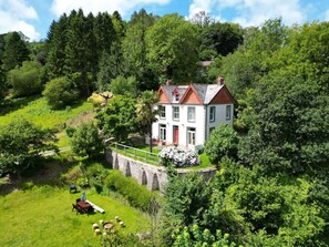 A two-story house with a red door, surrounded by lush greenery and a stone bridge. - The Woodlands House Estate (Berrynarbor)
