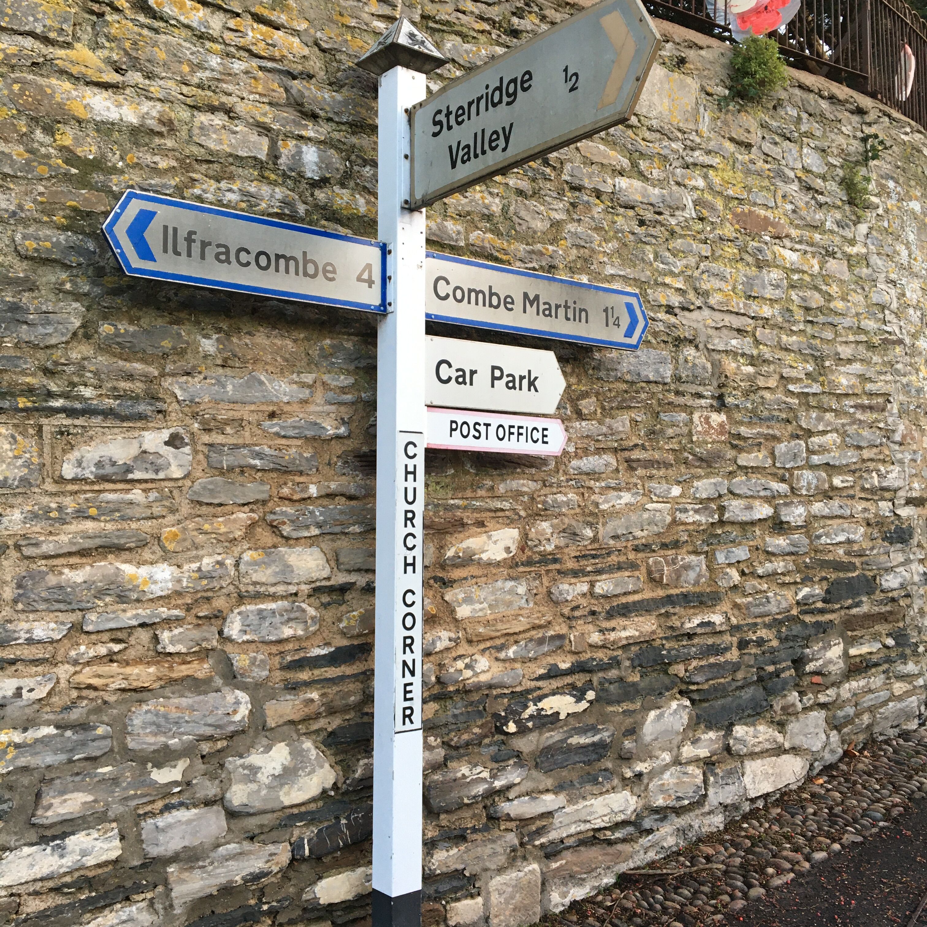 Um sinal de direção com várias setas indicando diferentes destinos, como Ilfracombe, Vale de Storridge, Combe Martin, Parque de Estacionamento, Correios e Igreja Corner.