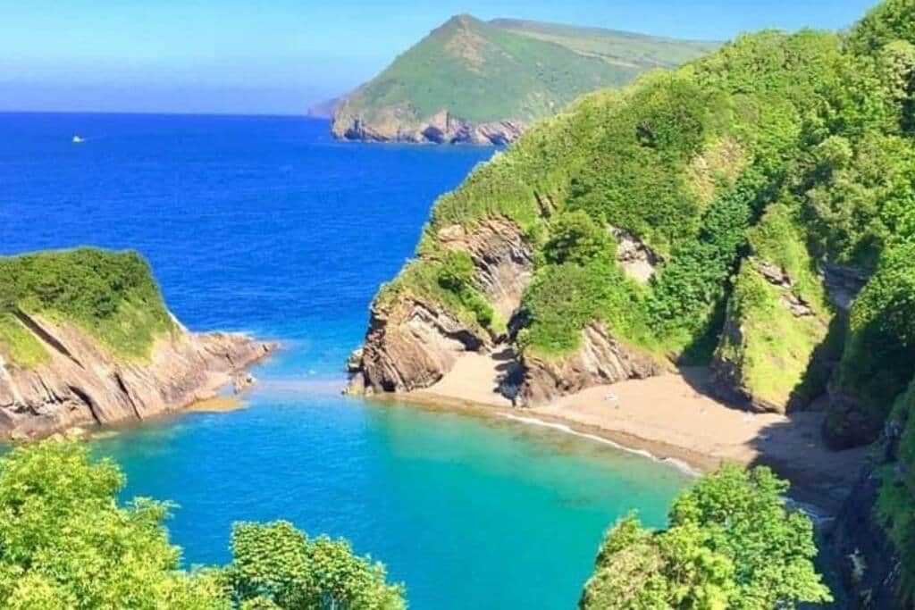 A coastal landscape with a narrow beach, rocky cliffs, and a clear blue sea.
