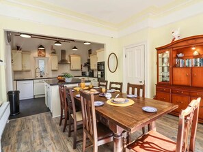 A dining area with a wooden table set for four, surrounded by chairs. A kitchen is visible in the background with modern appliances and cabinets.