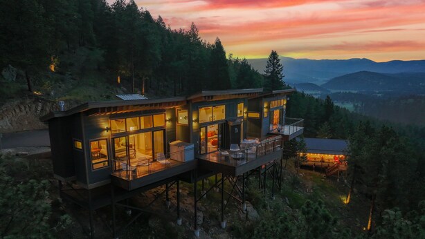 A modern, elevated cabin with large windows and a balcony, surrounded by forested hills.