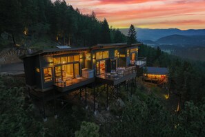 A modern, elevated cabin with large windows and a balcony, surrounded by forested hills. - Incredible Luxury Zen Treehouse+Glamping Tent - Views for Miles & Hot Tub/Sauna (Bailey)