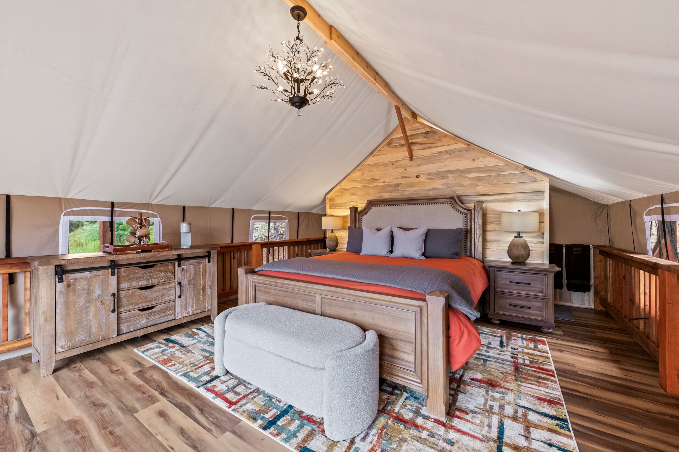A spacious tent with a wooden ceiling and a large bed with a red blanket. There is a bench and a small table with a lamp.