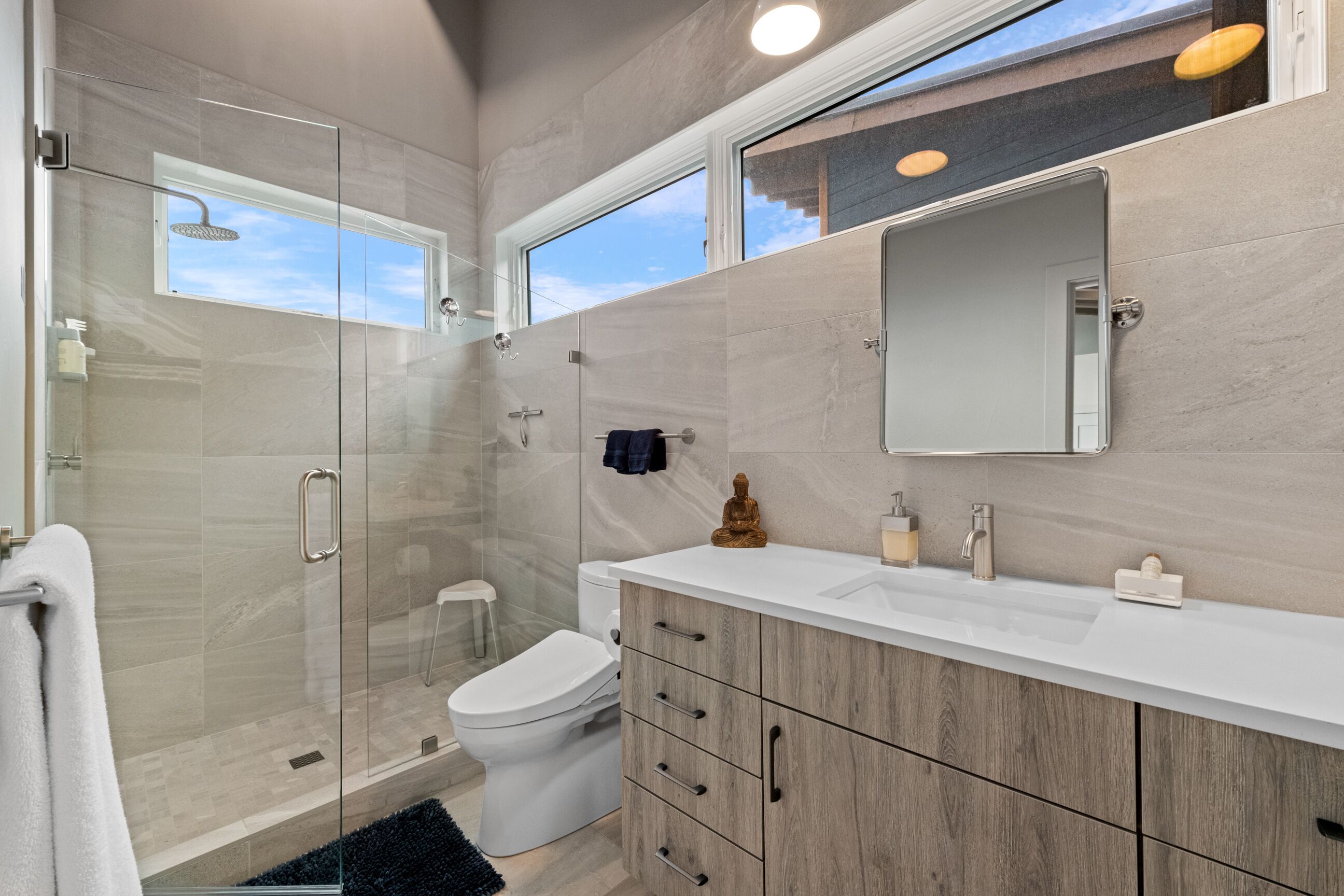 Een moderne badkamer met een douchecabine van glas, een wit toilet, een houten wastafelmeubel met spiegel, en een klein krukje.