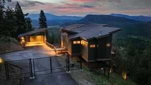 A modern two-story building with a flat roof, a covered outdoor area, and a view of mountains in the background.