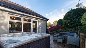 Uma área de spa com hidromassagem, um espaço de estar com mesa, e vista para um jardim com árvores e sebes.